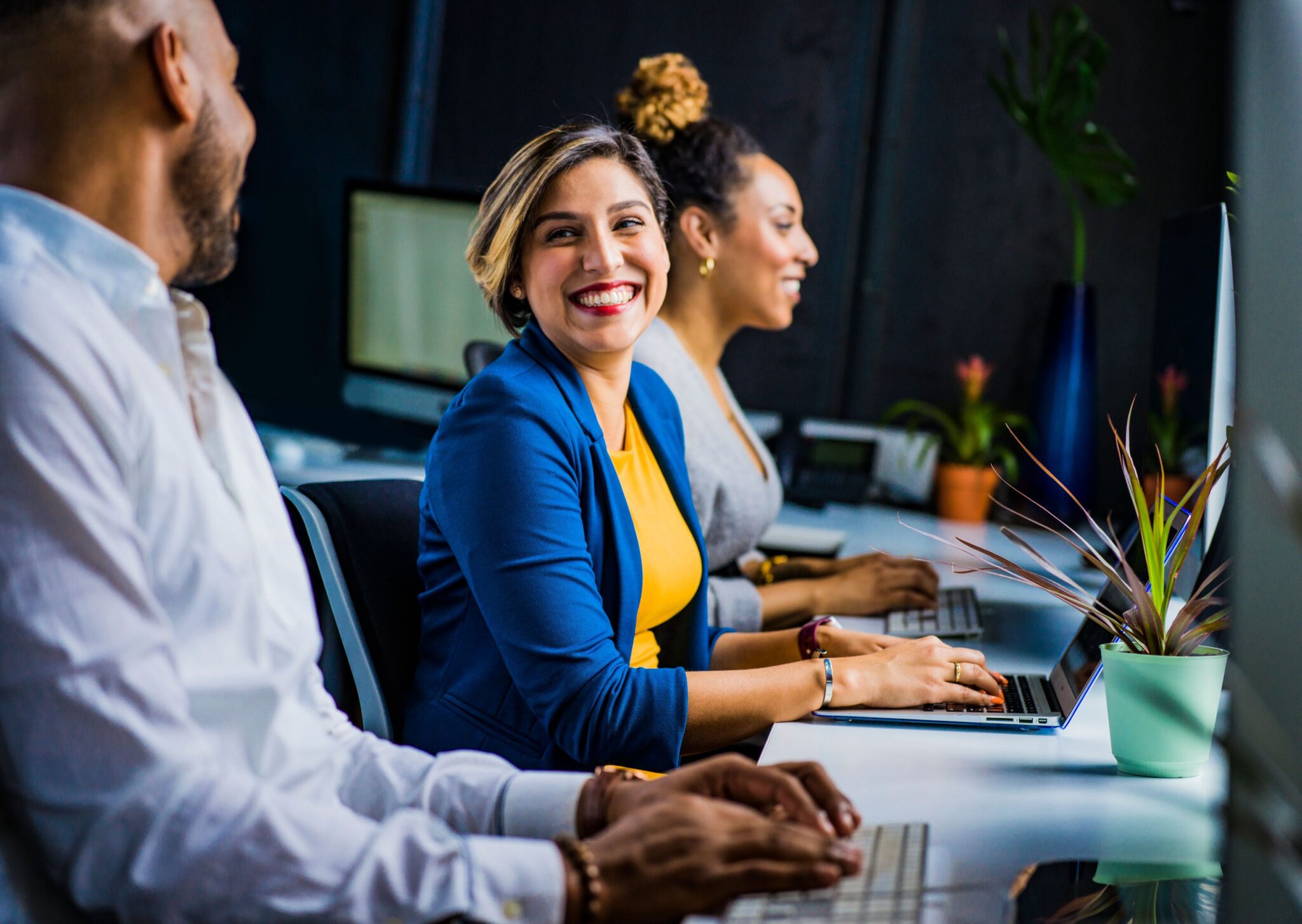 Smiling woman in a blue blazer and yellow top sits at a desk with a laptop in a modern office, flanked by two coworkers. Image Credit: Jopwell